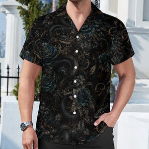 May include: A short-sleeved button-down shirt in black featuring a detailed print of dark teal roses, winding snakes, and intricate gold-toned filigree patterns. The shirt has a classic collar, a straight hem, and white buttons running down the front.
