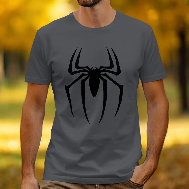 Puede incluir: Camiseta gris con un gran dise&ntilde;o de ara&ntilde;a negra en la parte delantera. El dise&ntilde;o de la ara&ntilde;a es una ilustraci&oacute;n sim&eacute;trica y estilizada con patas largas y curvas y un cuerpo central. La camiseta es de color s&oacute;lido y tiene cuello redondo.