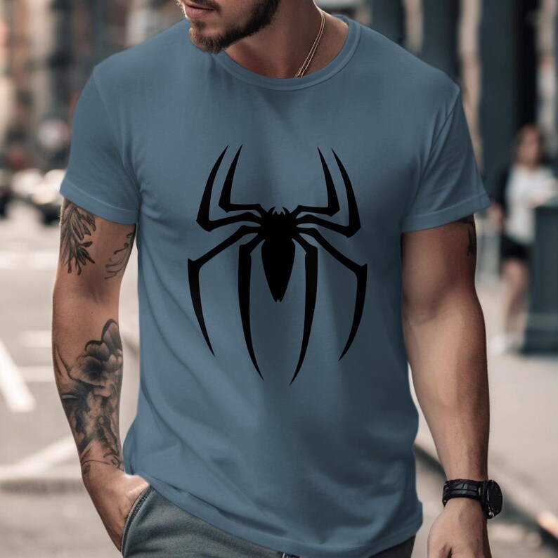 Puede incluir: Camiseta azul con un gran dise&ntilde;o de ara&ntilde;a negra en la parte delantera. El dise&ntilde;o de la ara&ntilde;a tiene patas largas y curvas y un cuerpo detallado. La camiseta tiene mangas cortas y cuello redondo.