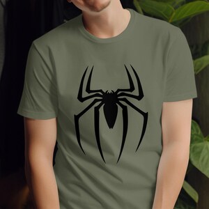 Puede incluir: Camiseta verde oliva con un gran dise&ntilde;o de ara&ntilde;a negra en la parte delantera. El dise&ntilde;o de la ara&ntilde;a tiene patas largas y angulares y un cuerpo detallado. La camiseta est&aacute; hecha de un material suave y tiene mangas cortas.
