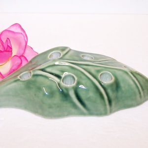 May include: A green ceramic leaf-shaped flower frog with multiple holes for arranging stems. The frog has a glossy finish and is designed to hold flowers. A pink flower is in the background.
