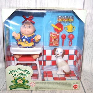 May include: A Cabbage Patch Kids doll set in a clear plastic box. The set includes a doll in a high chair, a toy dog, and miniature accessories like food jars, a spoon, and a baby bottle. The box has a red and white checkered background.