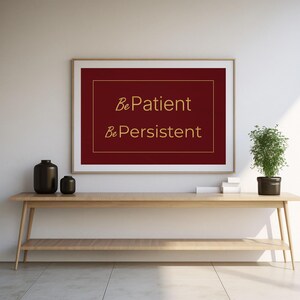 Puede incluir: Un cuadro enmarcado con las palabras "Be Patient, Be Persistent" en letras doradas sobre un fondo burdeos. El cuadro cuelga sobre una consola de madera con jarrones negros y una planta en maceta.