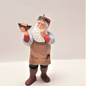 May include: A Santa Claus figurine wearing a brown apron and holding a small wooden boat. He is wearing a red and white striped shirt, a green hat with a red band, and brown boots.