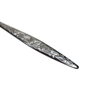 May include: Close-up of a silver utensil with a textured, woven-like pattern on the handle. The utensil has a pointed end and a polished, reflective surface, suggesting it is made of metal.