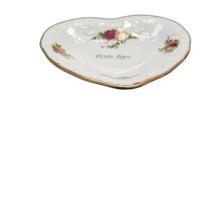 May include: Heart-shaped, white ceramic dish with a gold rim. The dish features floral accents in pink, red, and green, with the words "With love" printed in the center. A decorative piece for trinkets or small items.