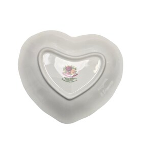 May include: White heart-shaped dish with a floral design in the center. The dish has a smooth, glossy finish and is made of ceramic. The floral design includes pink, red, and purple roses. The dish is from the Royal Albert brand.