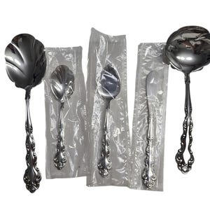 May include: Four silver serving pieces in individual plastic packaging. The pieces include a butter knife, a small spoon, a larger spoon, and a serving spoon with a scalloped edge.