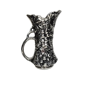 May include: A silver-colored pitcher with a handle and a detailed grape and leaf design. The pitcher has a flared top and a round base. The handle is attached to the side of the pitcher with a decorative loop.