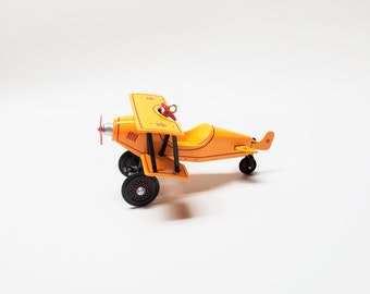 May include: A vintage-style toy airplane in bright yellow with red accents. The biplane features a propeller, wheels, and a star detail on the tail. The toy is set against a white background.
