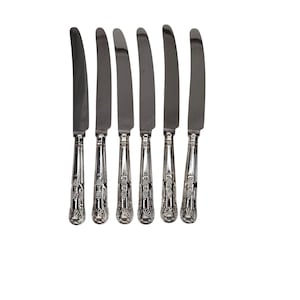 May include: Six silver butter knives with ornate handles. The blades are curved and the handles feature a detailed floral and shell design. The knives are arranged in a row against a white background, showcasing their elegant design.