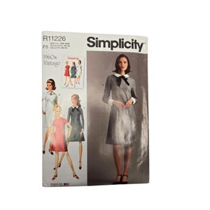 May include: A Simplicity sewing pattern envelope R11226 for a 1960s vintage dress. The front shows a gray dress with a white collar, white cuffs, and a black bow tie. Illustrations of the dress in various colors and styles are also displayed.