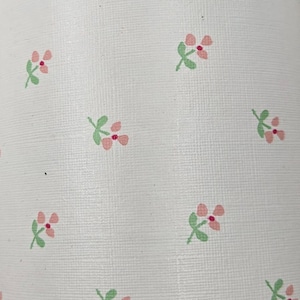 May include: White fabric with a repeating pattern of small pink flowers and green leaves. The flowers have three petals and are evenly spaced across the surface. The fabric has a slightly textured appearance.