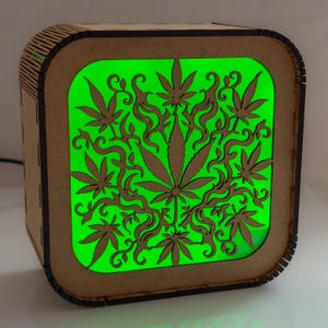 Puede incluir: Una lámpara cuadrada de madera con esquinas redondeadas y un panel frontal cortado con láser. El panel muestra un patrón detallado de varias hojas de cannabis y tallos en forma de enredadera. El interior emite una luz verde brillante.