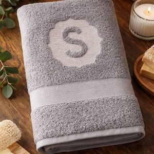 Peut inclure: Une serviette de bain grise avec la lettre "S" brodée dans un motif décoratif blanc. La serviette est pliée et a une surface texturée. La serviette est sur une surface en bois, avec une bougie et du savon à proximité.