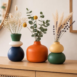 May include: Three ceramic vases in distinct shapes and colors. The first is a stacked dark blue, white, and green vase with dried grasses. The second is a round orange vase with eucalyptus. The third is a stacked yellow and green vase with lavender.