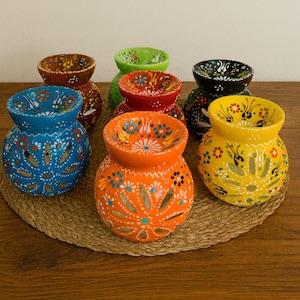 May include: A collection of hand-painted ceramic vases in various colors, including blue, orange, and yellow. Each vase features a floral design and sits on a woven mat. The vases are decorative.