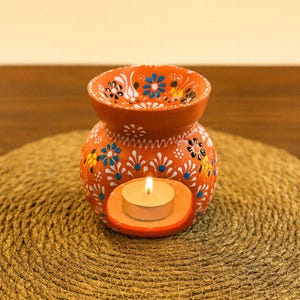 Puede incluir: Un quemador de aceite de terracota con un patrón floral pintado a mano en azul, blanco y amarillo. El recipiente redondo tiene un cuenco superior ancho y una abertura arqueada inferior que contiene una pequeña vela blanca encendida.