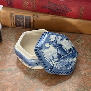 May include: A hexagonal ceramic trinket box featuring a blue and white hand-painted scene of a windmill on the lid. The base is white with blue decorative accents along the edges. The lid is partially removed, revealing the hollow interior.
