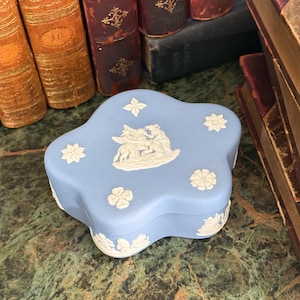 May include: A light blue, flower-shaped trinket box featuring white relief ornamentation. The lid displays a central mythological scene with a winged figure and surrounding floral motifs, while the sides are decorated with additional white relief figures.