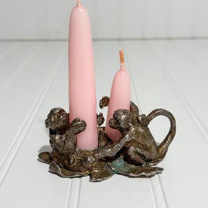 May include: A vintage candle holder with a bronze finish, featuring three monkey figures. Two pink taper candles are inserted into the holder. The candle holder is placed on a white surface.