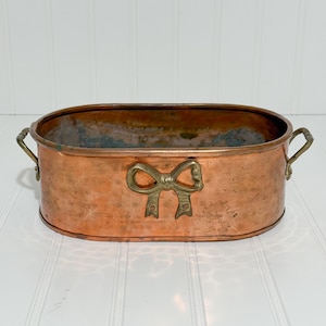 May include: An oval copper planter with brass handles and a brass bow accent. The planter has a weathered patina, suggesting age and use. The handles are small and curved, and the bow is centered on the front.
