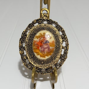 May include: An oval-shaped, gold-toned pendant with a detailed filigree border. The center features a painted scene of two figures in a landscape. The pendant is displayed on a small, adjustable easel.