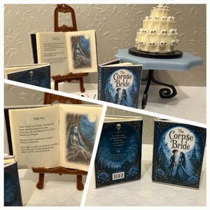 以下が含まれることがあります： 青い表紙のミニチュア本「The Corpse Bride」が茶色のイーゼルに飾られています。開いたページには印刷されたテキストと女性キャラクターのイラストが描かれています。小さな頭蓋骨で飾られた白いケーキが青いスタンドの上に置かれています。