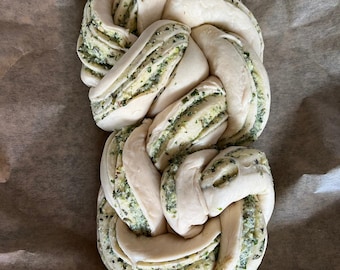 May include: A braided loaf of bread with visible layers of fresh herbs, garlic, and Parmesan cheese. The bread is light in color with green flecks from the herbs. The text "Fresh Herb, Garlic & Parmesan Cheese Twist" is at the top.