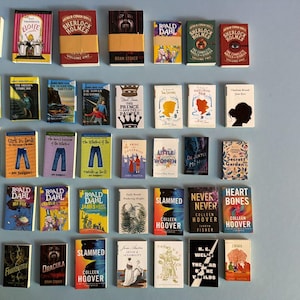 May include: A selection of miniature books with diverse covers and titles, such as "Sherlock Holmes," "Moby Dick," and "Slammed." The books are displayed on a light blue surface, featuring a range of colours and designs.
