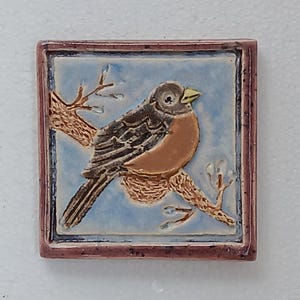 May include: Square ceramic tile featuring a detailed illustration of a robin perched on a branch. The bird has brown and orange feathers, a yellow beak, and is set against a light blue background. The tile is framed with a dark red border.
