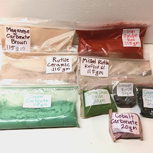 May include: A collection of eight clear plastic bags containing various powdered pigments. Labels include Manganese Carbonate Brown 115gm, Iron Oxide Red 115gm, Rutile Ceramic 115gm, Milled Rutile Reflux 61 115gm, Copper Carbonate 115gm, Chrome Green Oxide 20gm, Granular Rutile 20gm, Copper Oxide Black 20gm, and Cobalt Carbonate 20gm.