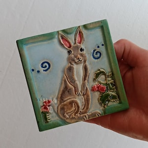 May include: Square ceramic tile featuring a detailed relief of a standing rabbit with pink-lined ears, set against a light blue background. The tile has a green border and includes stylized floral and swirl designs. The tile measures approximately 4 inches square.