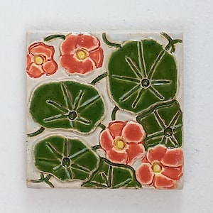 May include: Square ceramic tile featuring a floral design. The tile showcases red flowers with yellow centers and green leaves with white veins. The background is a light cream color, and the tile has a glossy finish.
