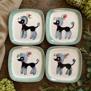May include: A set of four square ceramic plates with rounded corners, each featuring a hand-painted black poodle with grey fur accents, a red collar, and a small pink flower above its head, framed by a speckled light green border.