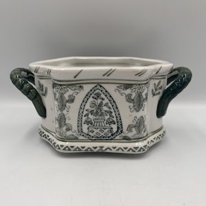 Peut inclure: Un cache-pot en céramique blanche de forme hexagonale, orné de motifs floraux gris foncé et d'un motif central en arche représentant une plante en pot. Le récipient possède deux anses latérales texturées gris foncé.