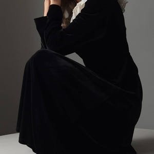 May include: A long, black velvet dress with a white lace collar. The dress has long sleeves and a flowing skirt. The model is seated, with hands near the face. The dress is a classic, elegant style.