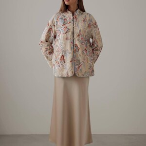 May include: A beige, floral-patterned jacket with a mandarin collar and matching skirt. The jacket features a colorful floral design with red, blue, and pink accents. The skirt is a solid, neutral beige color. The outfit is completed with beige heels.