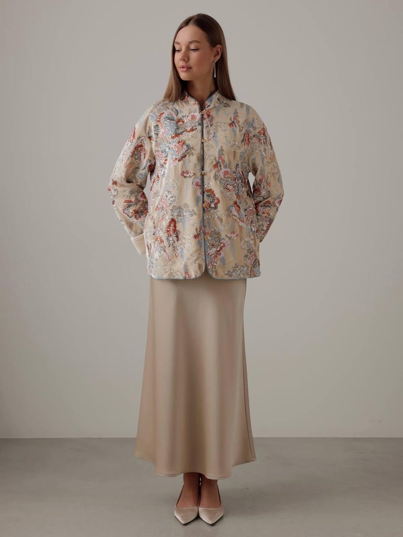 May include: A beige, floral-patterned jacket with a mandarin collar and matching skirt. The jacket features a colorful floral design with red, blue, and pink accents. The skirt is a solid, neutral beige color. The outfit is completed with beige heels.