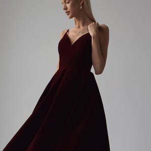 May include: A burgundy-colored formal gown with a V-neckline and spaghetti straps. The dress has a full skirt and is made of a soft, flowing fabric. The model is wearing the dress in a studio setting.