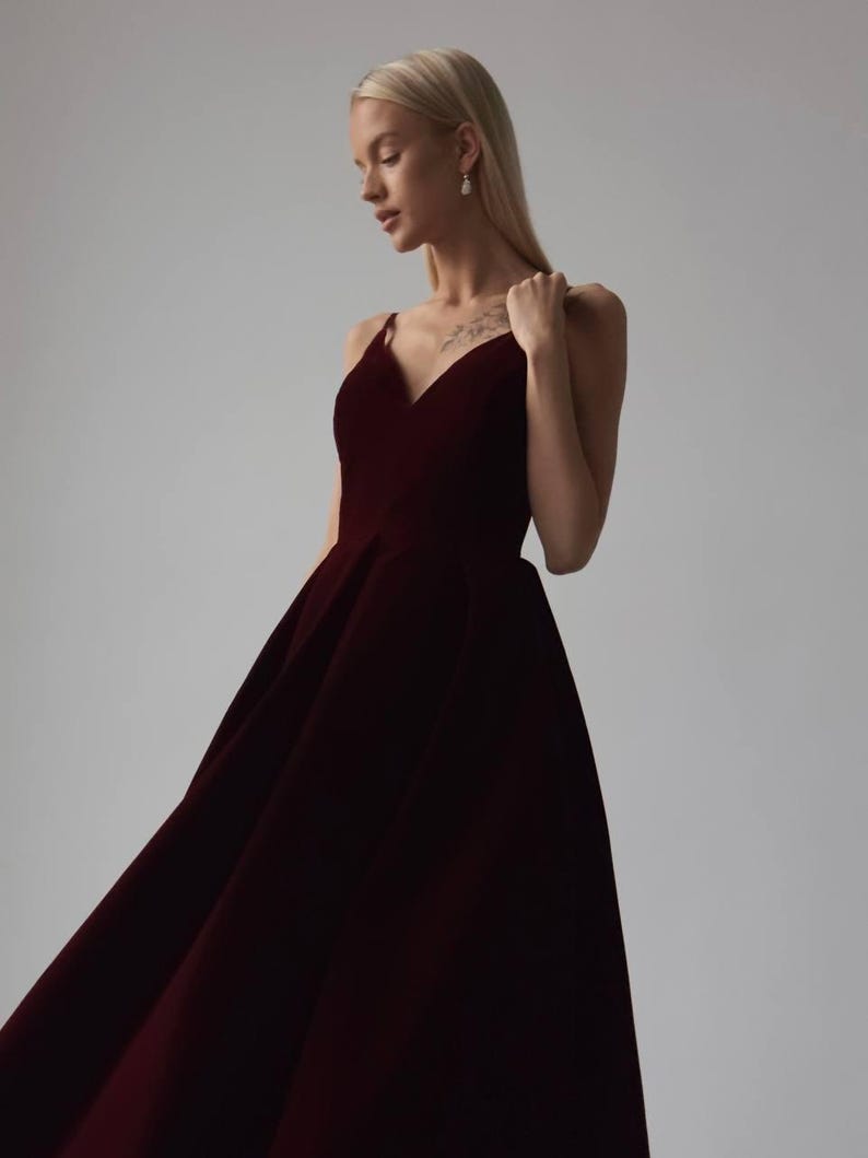 May include: A burgundy-colored formal gown with a V-neckline and spaghetti straps. The dress has a full skirt and is made of a soft, flowing fabric. The model is wearing the dress in a studio setting.