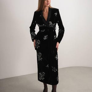 May include: A black velvet wrap dress with floral embroidery. The dress features long sleeves, a belted waist, and a shawl collar. The floral embroidery is silver and white. The dress is knee-length.