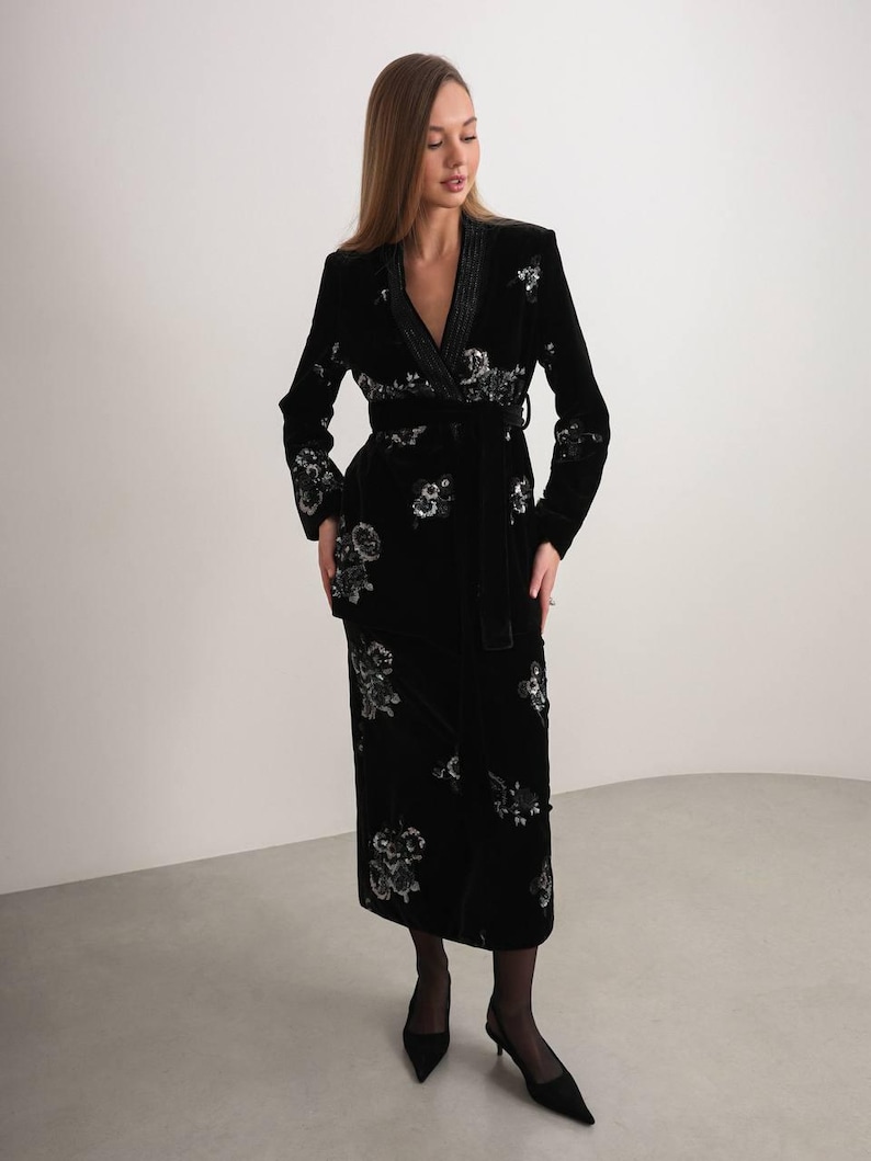 May include: A black velvet wrap dress with floral embroidery. The dress features long sleeves, a belted waist, and a shawl collar. The floral embroidery is silver and white. The dress is knee-length.