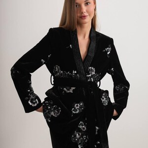 May include: A black velvet robe with silver sequin floral embroidery. The robe features a shawl collar, a matching belt, and pockets. The sleeves and front of the robe are adorned with the floral design. The robe is a luxurious piece of clothing.