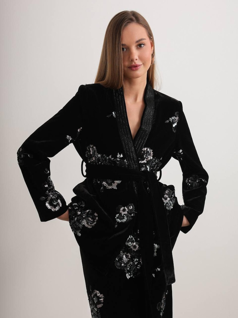 May include: A black velvet robe with silver sequin floral embroidery. The robe features a shawl collar, a matching belt, and pockets. The sleeves and front of the robe are adorned with the floral design. The robe is a luxurious piece of clothing.