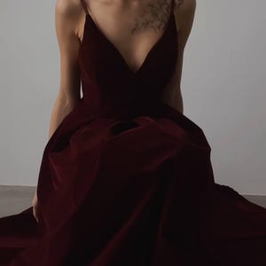 May include: A deep burgundy velvet gown with a plunging neckline and spaghetti straps. The dress has a full skirt and appears to be floor-length. The fabric has a rich, luxurious appearance, suggesting a formal occasion.