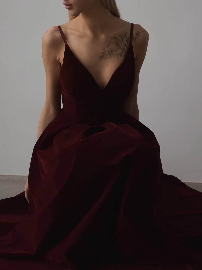 May include: A deep burgundy velvet gown with a plunging neckline and spaghetti straps. The dress has a full skirt and appears to be floor-length. The fabric has a rich, luxurious appearance, suggesting a formal occasion.