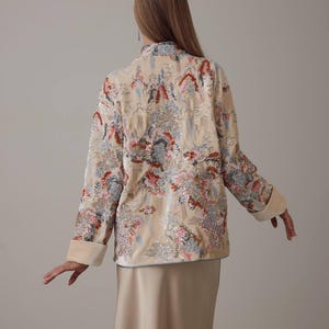 May include: A beige jacket with a floral pattern in shades of red, blue, and pink. The jacket has a mandarin collar, long sleeves with turned-up cuffs, and a satin-like finish. The model is wearing a matching beige skirt.