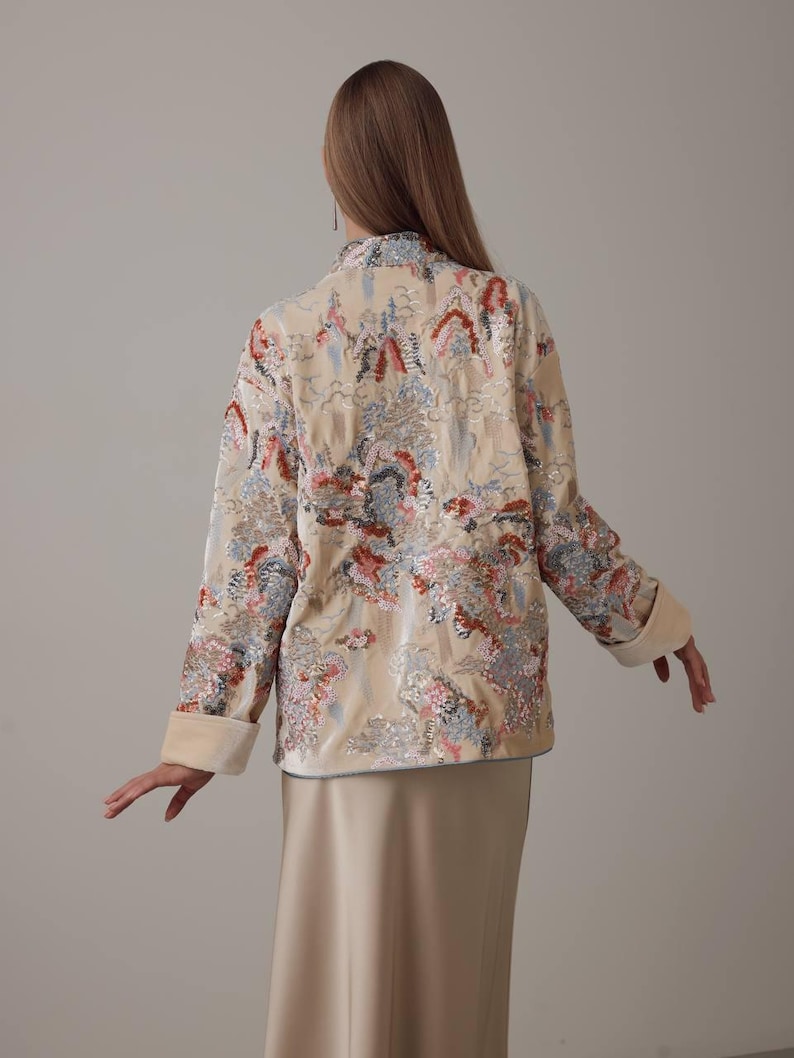May include: A beige jacket with a floral pattern in shades of red, blue, and pink. The jacket has a mandarin collar, long sleeves with turned-up cuffs, and a satin-like finish. The model is wearing a matching beige skirt.