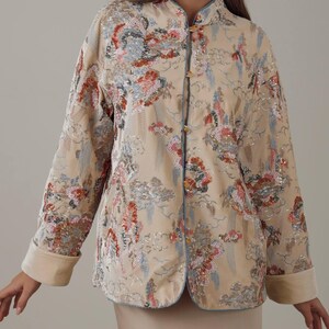 May include: Beige jacket with floral embroidery and sequin details. The jacket features a mandarin collar, long sleeves with rolled cuffs, and gold-colored buttons. The design includes blue trim and a subtle pattern.